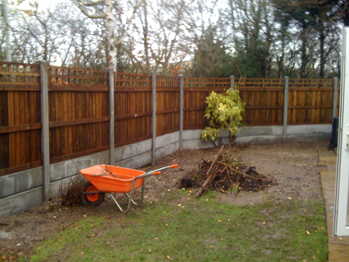 fencing surrounding a garden with a small island of dirt next to an orange wheelbarrow